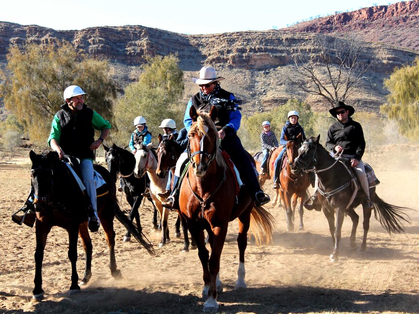 A group of horse riders start their journey. A large rocky mountain range is in the background.