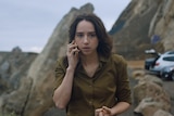 White woman with mid-length brunette hair wears olive green shirt and talks on a mobile phone outside near the ocean.