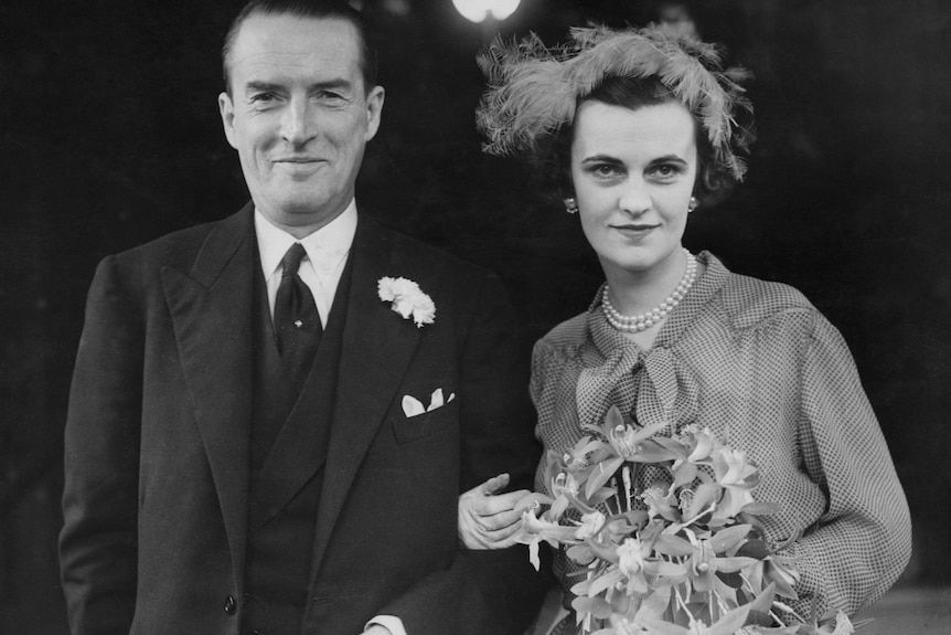 A man wearing a dark suit smiles with his arm around a woman wearing a dress and feather fascinator.