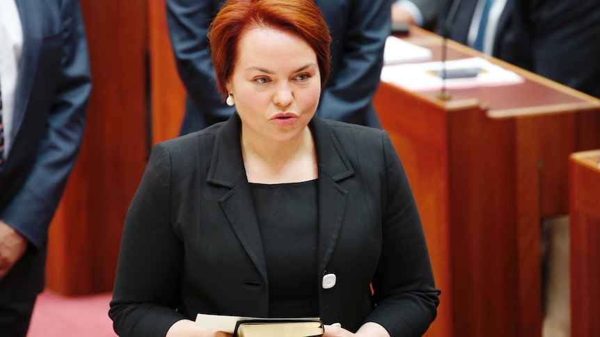 Woman (Kimberly Kitching) stands in senate holding a book. 