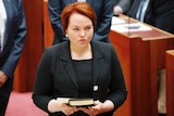 Woman (Kimberly Kitching) stands in senate holding a book. 