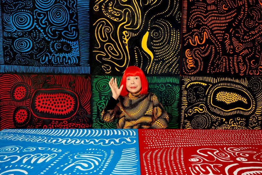 A 95-year-old Japanese woman in a black and yellow patterned top and with vibrant red hair, sits among large colourful prints.
