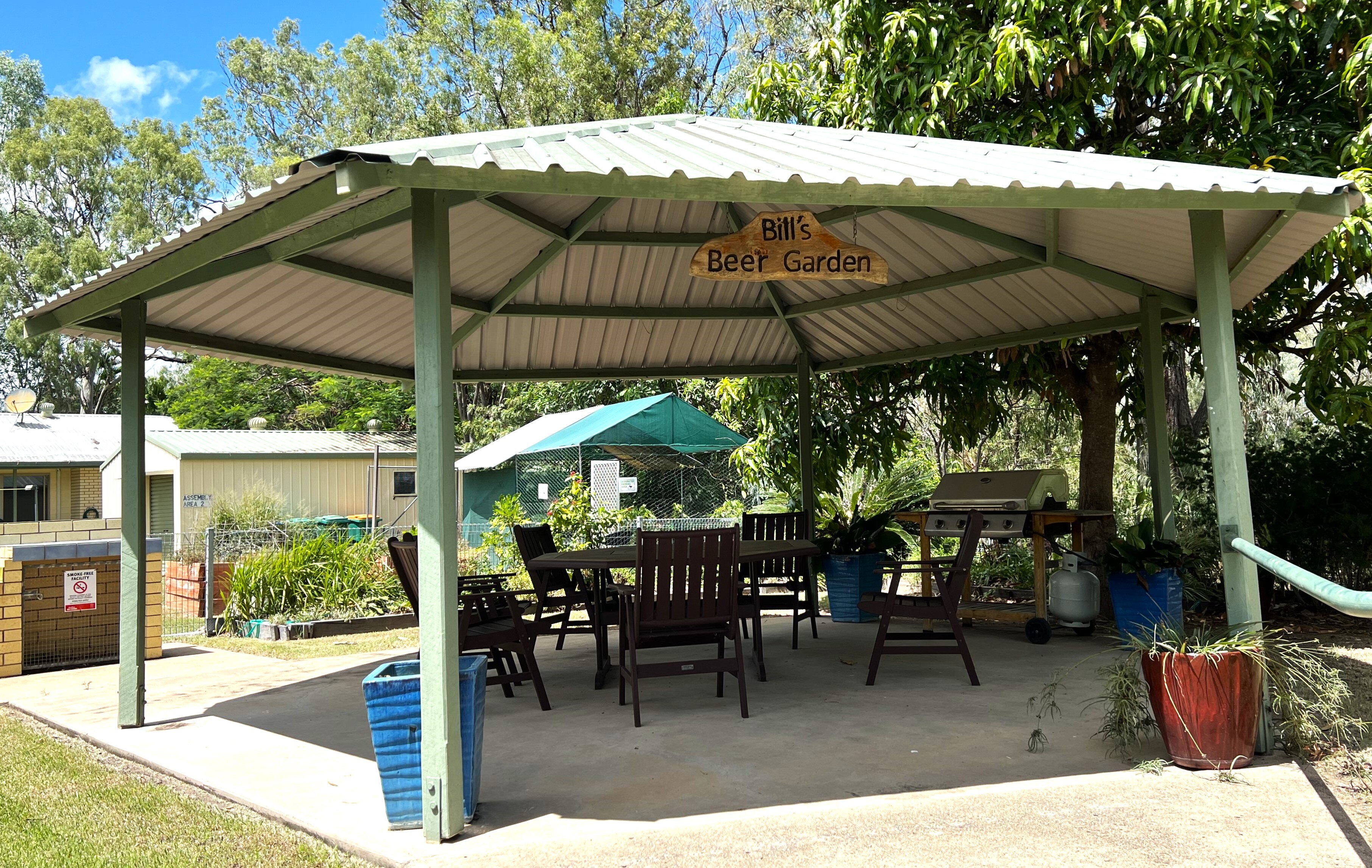 A patio under a pergola with a bbq and table and chairs, a sign reading Bill's Beer Garden hangs from ceiling