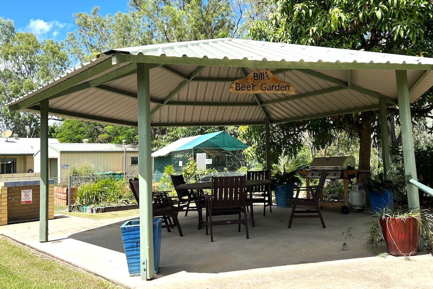 A patio under a pergola with a bbq and table and chairs, a sign reading Bill's Beer Garden hangs from ceiling