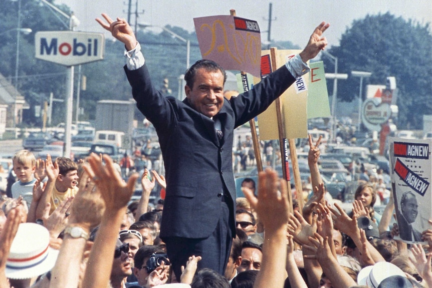 Richard Nixon stands among a crowd outdoors, giving his gives his trademark "victory" sign with fingers forming a V shape.