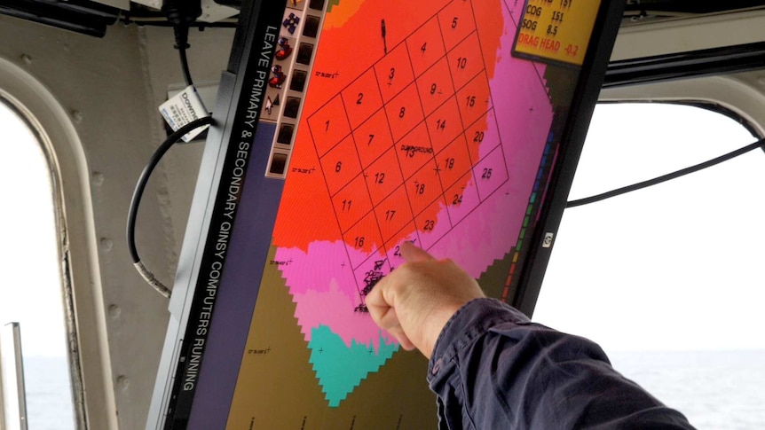 A man's hand points out a location on a computer screen showing an area on a map divided into grid locations