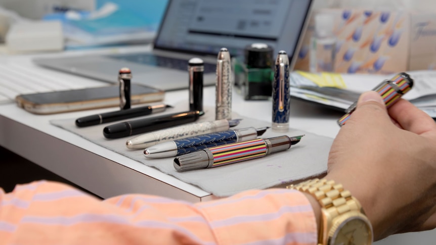 A row of fountain pens in various styles and colours sits on a desk.
