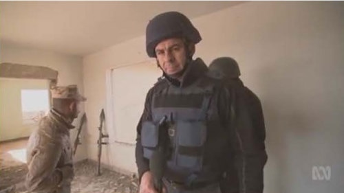 A journalist in an empty room, dressed in a bullet-proof vest and helmet, reports on the war with guns leaning against the wall.