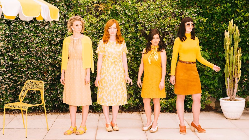 Four women dressed in retro-looking yellow clothes stand in front of a hedge near a large yellow umbrella and a yellow chair.