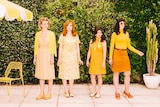 Four women dressed in retro-looking yellow clothes stand in front of a hedge near a large yellow umbrella and a yellow chair.