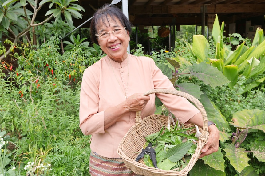 Woman wearing a light pink shirt smiles holding a basket of green produce in her garden.