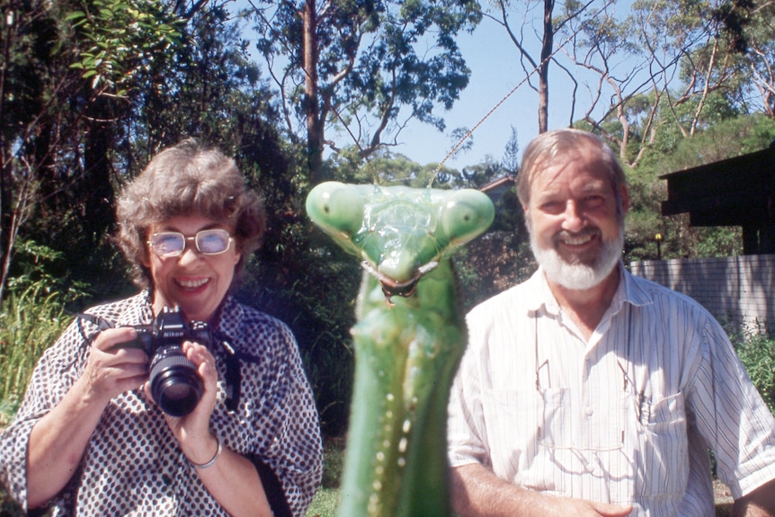 A woman is holding a camera and is sitting next to a man with a close-up of an insect in the foreground.