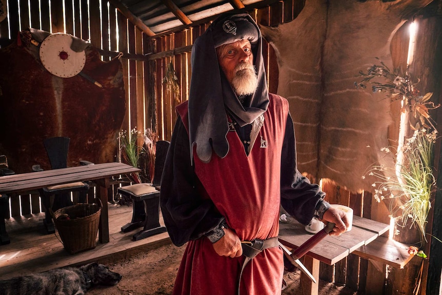 A man dressed in medieval red and blue clothes stands in a old style hall with a dog laying at his feet.