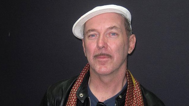 A man wearing a white cap and scarf standing against a dark grey wall.