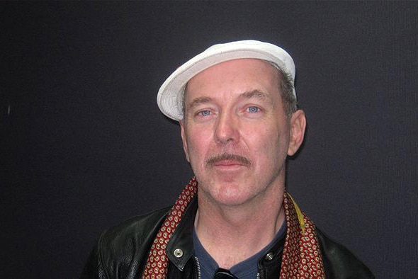 A man wearing a white cap and scarf standing against a dark grey wall.