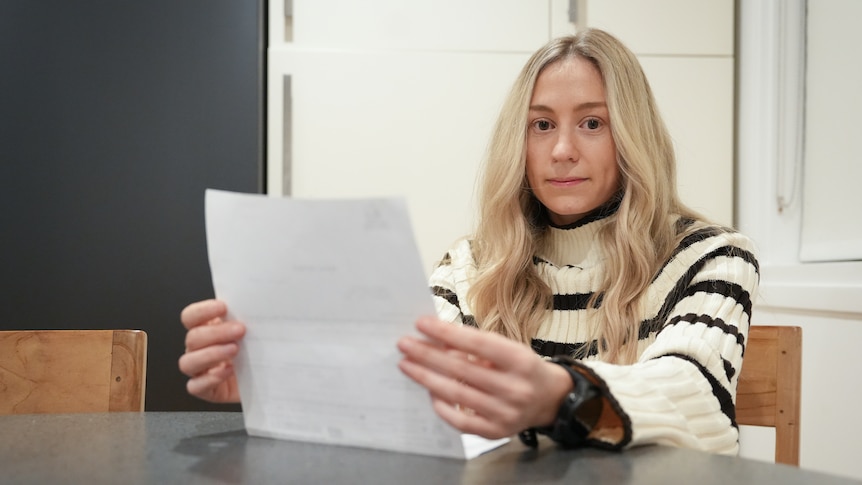 A woman holds a piece of paper and stares blankly while sitting at a dining table.