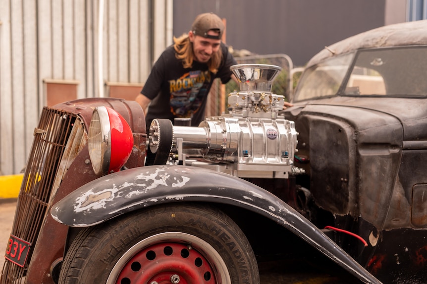 A smiling young man inspects a hot rod that has a burnt exterior.