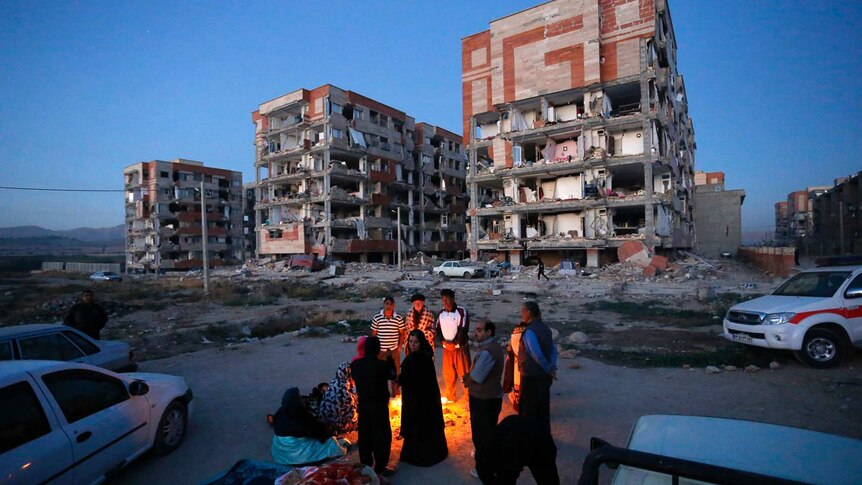 People stand around a fire in the foreground of a destroyed building.