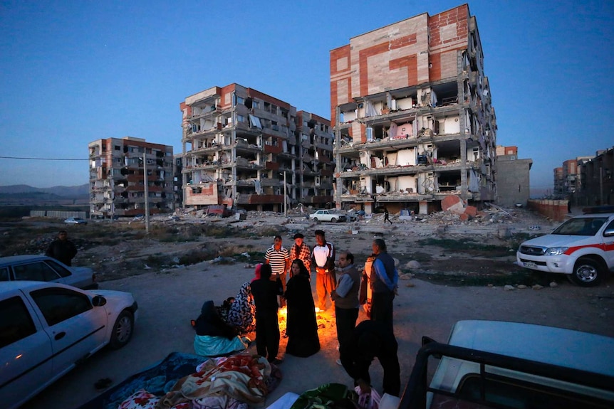 People stand around a fire in the foreground of a destroyed building.