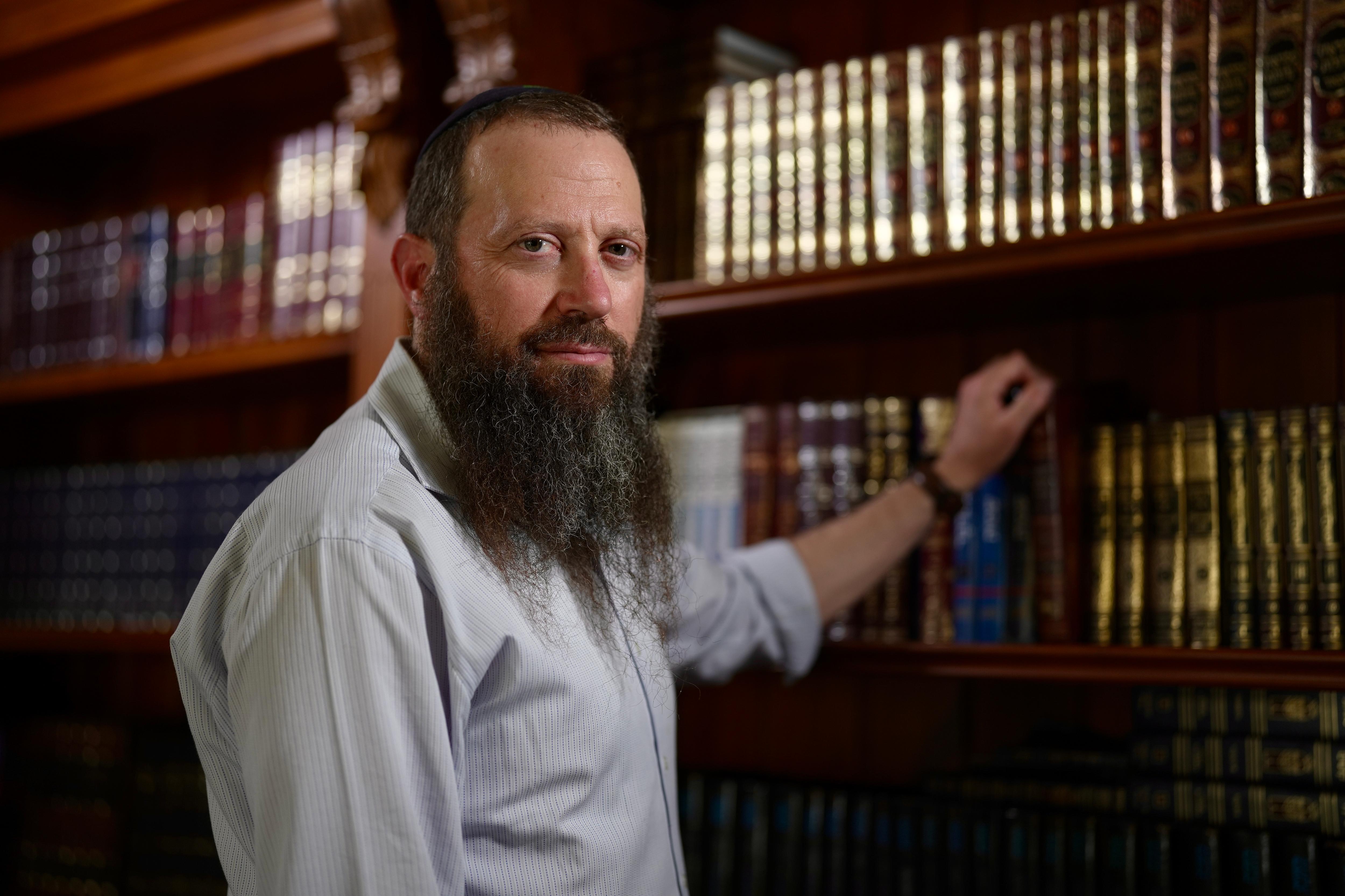 Um homem com uma longa barba escura veste uma camisa cinza de botão e fica parado com uma das mãos apoiada em uma estante, olhando para a câmera.