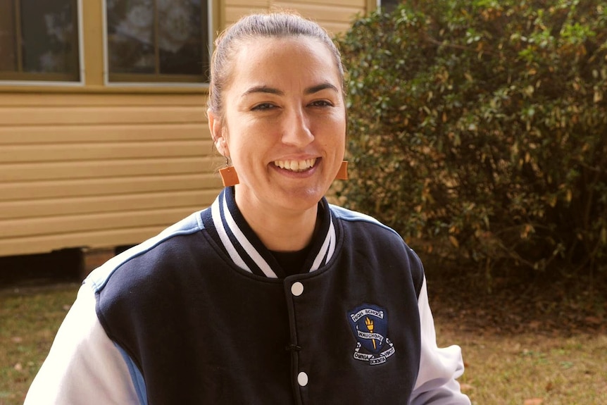 A woman in a sports jacket with the school emblem smiling to the camera with yellow weatherboard walls behind her.