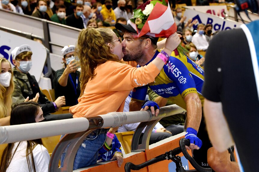 A cyclist on a track bike leans over a railing to kiss his daughter before a big race.