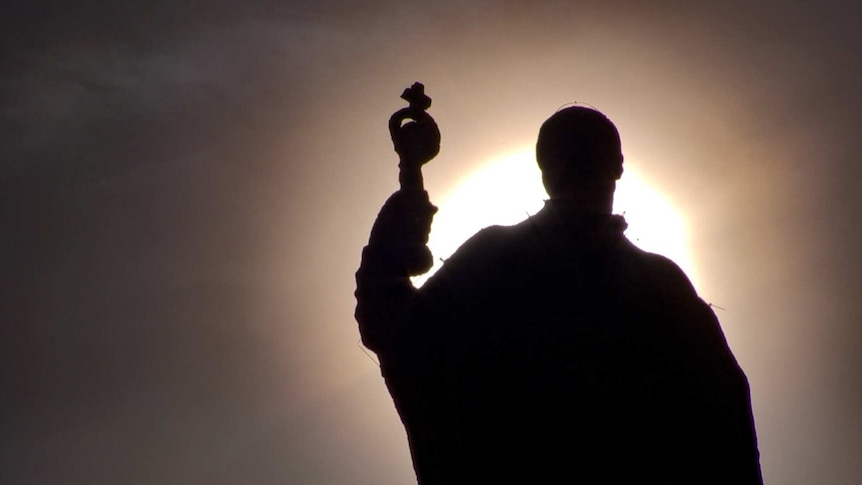 The sun behind a darkened statue of a man holding a religious cross.
