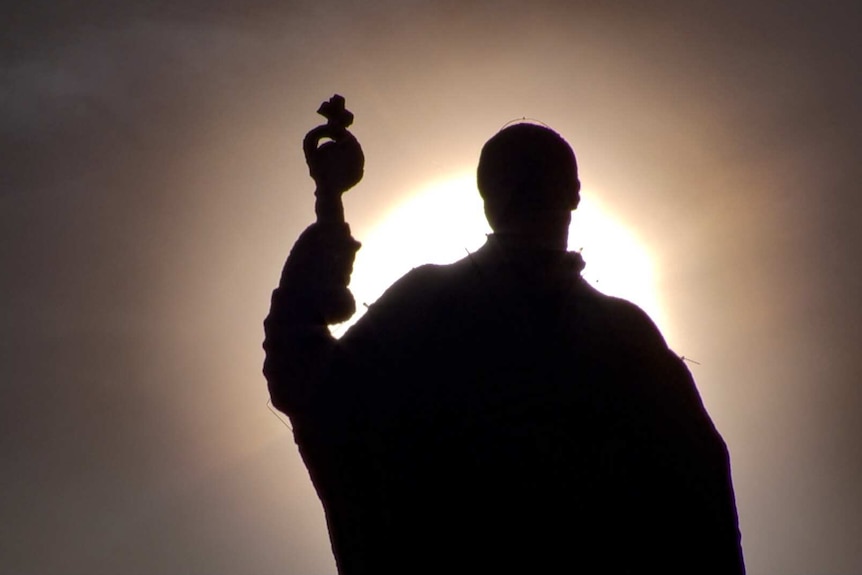 The sun behind a darkened statue of a man holding a religious cross.