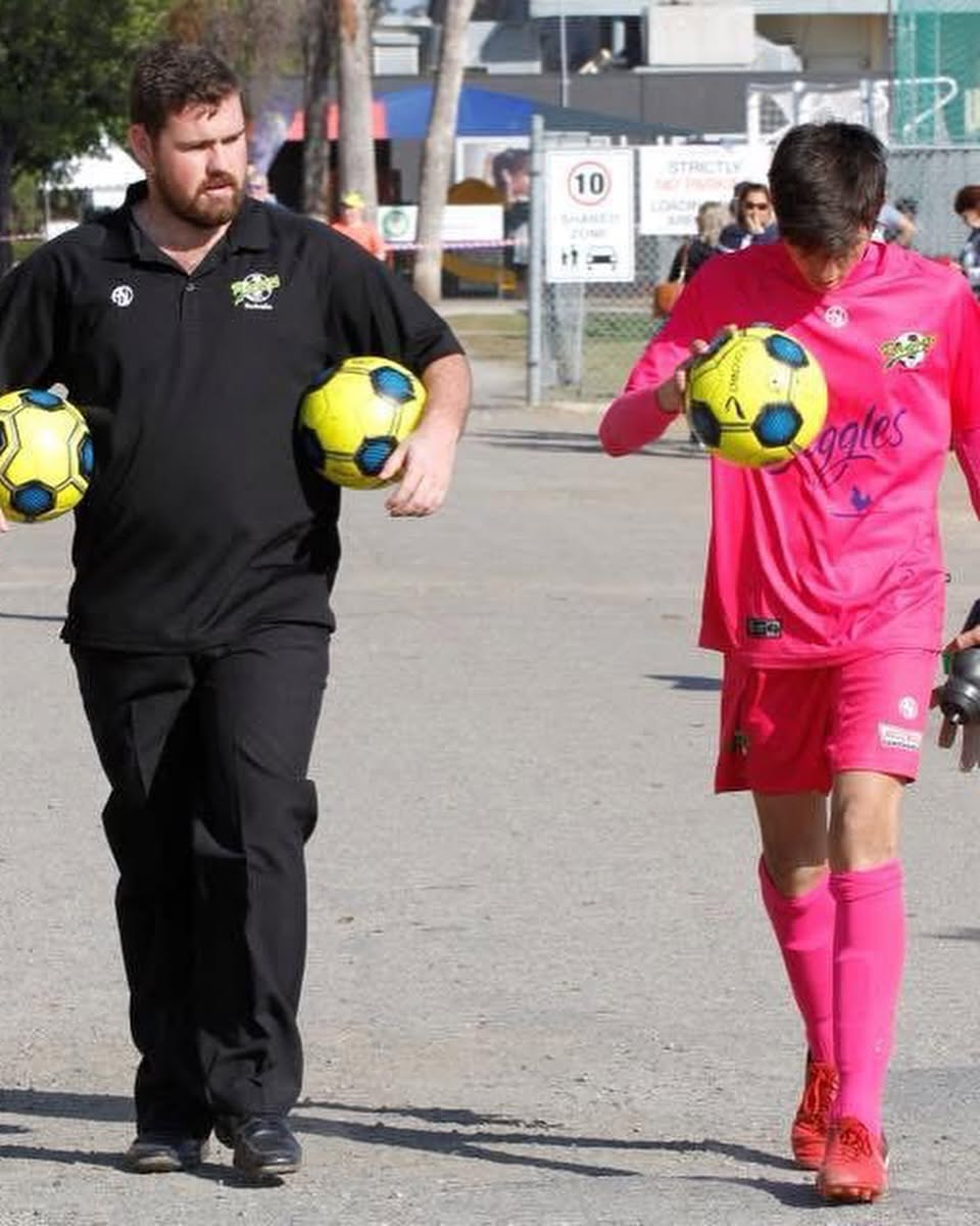 Mr Allen on the left carrying a soccer ball under each arm walking in a car park next to Beach also holding a ball.