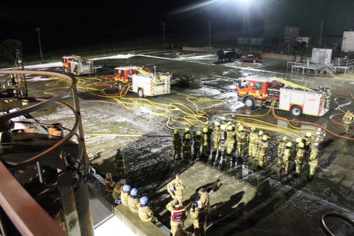A group of firefighters in yellow uniforms on a concrete training area with foam and water on the ground