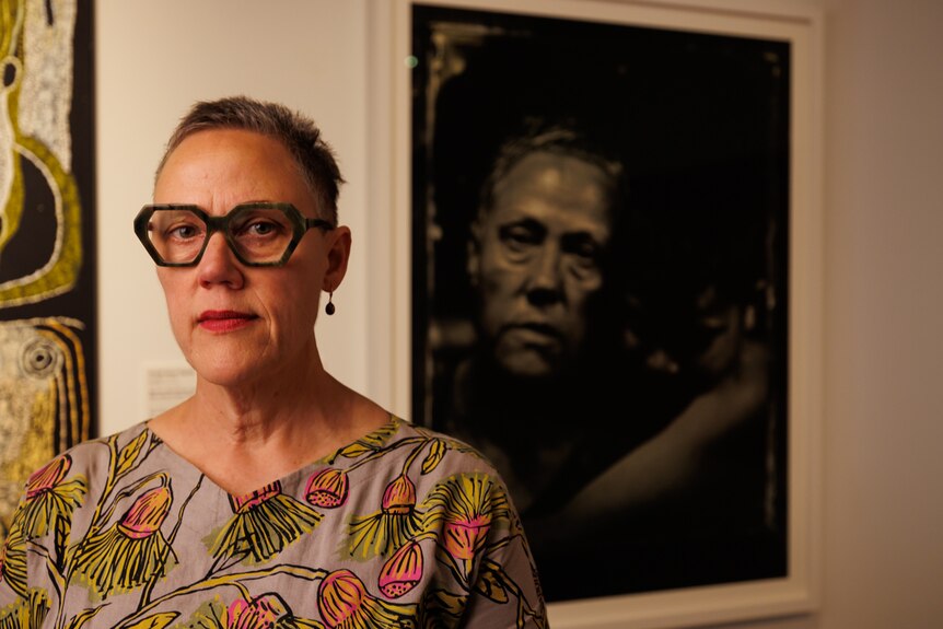 A woman wearing green glasses stands in front of a black and white photograph on a gallery wall