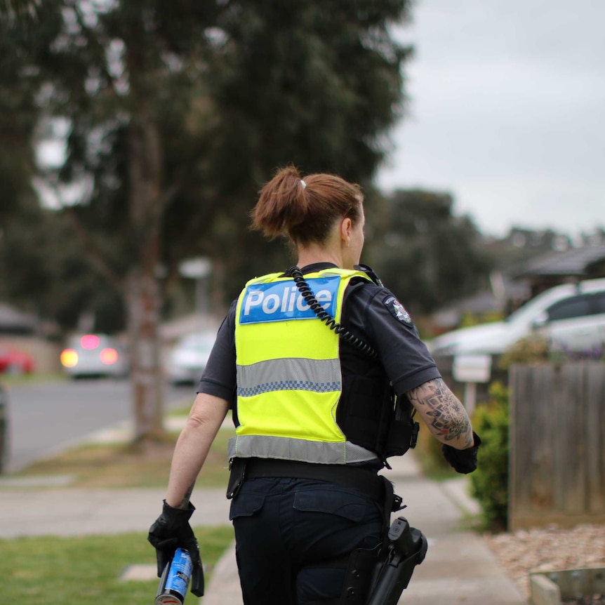 A female police officer is shown from behind walking down a suburban street, carrying pepper spray.