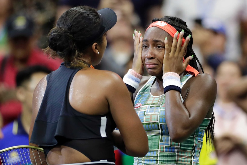 A tennis player wipes her eyes as she stands at the net talking to the player who beat her.