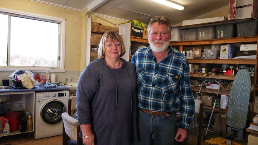 An older couple stand in what looks like a shed, their arms around each other. They look a bit beleaguered.