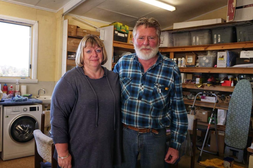 An older couple stand in what looks like a shed, their arms around each other. They look a bit beleaguered.