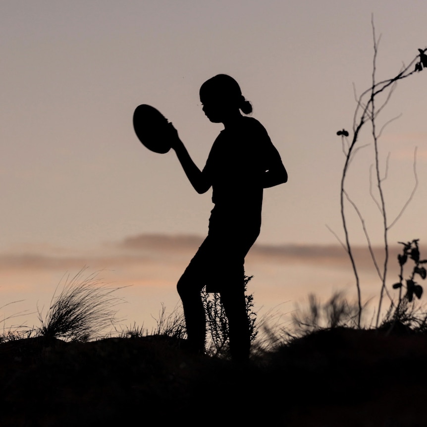 A young girl is silhouetted against fading evening sun holding a football in one hand while walking across sand dune ridge