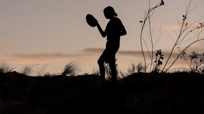 A young girl is silhouetted against fading evening sun holding a football in one hand while walking across sand dune ridge