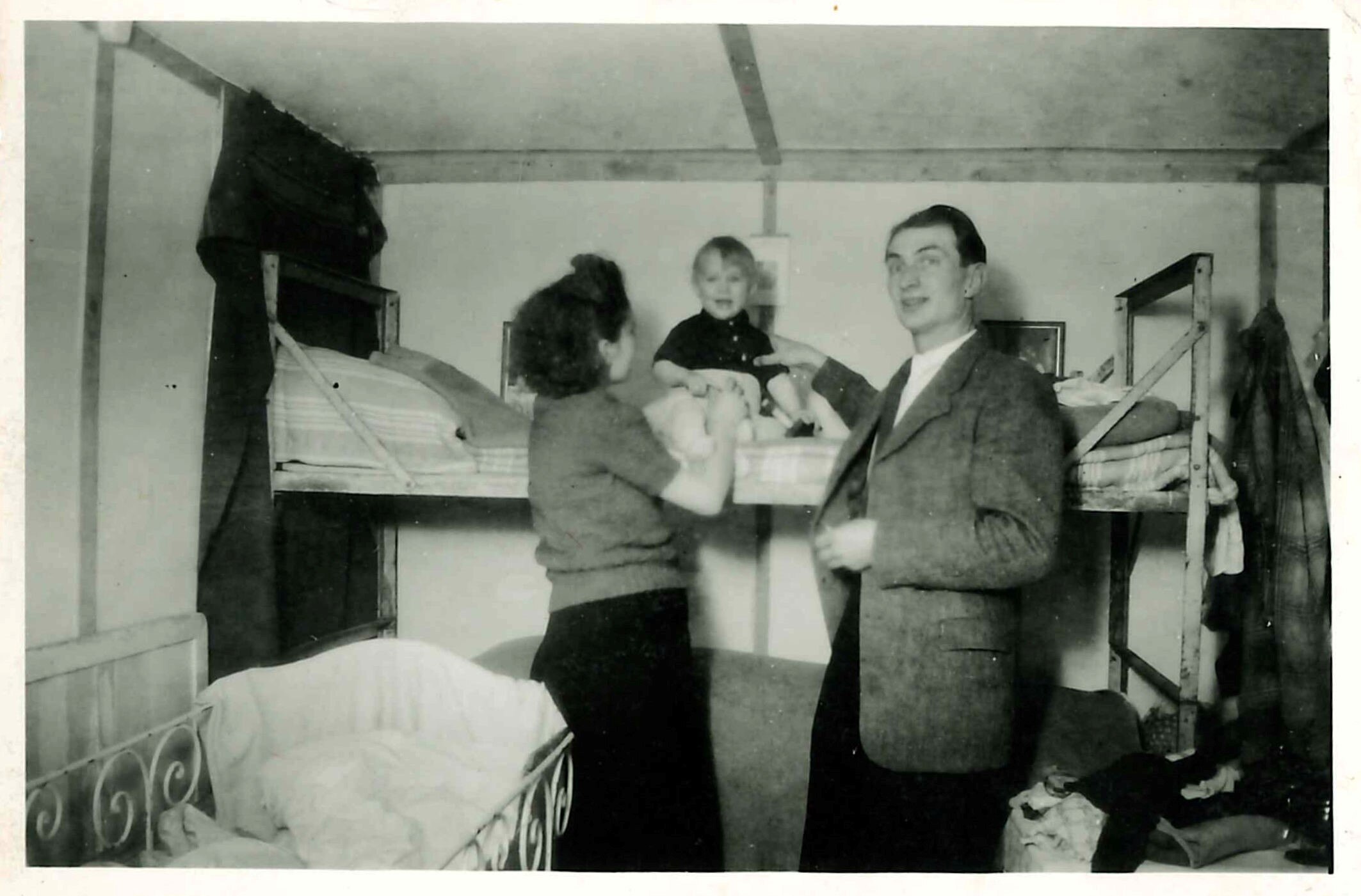 Woman and man stand next to bunk bed. Laughing child sits on bed. Flat is very small and basic.