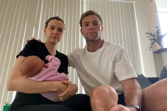 A woman and a man sit on a couch posing for a photo, with the woman holding a young baby in her arms.