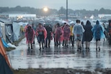 Festival goers wear ponchos and hold drinks as they walk in a group with grey skies above