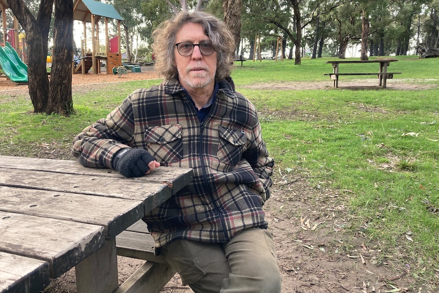 Sudhir wears a brown flannelette shirt, glass and dark fingerless gloves and sits at a park bench in a park near a playground