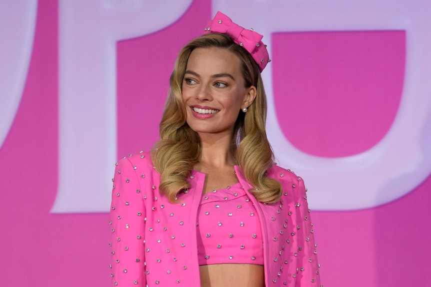 Margot Robbie wears a pink jacket and headpiece in front of a pink and white background.