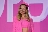 Margot Robbie wears a pink jacket and headpiece in front of a pink and white background.