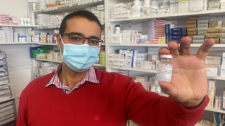 A man in a red jumper wearing glasses and holding a small glass bottle of vaccine 