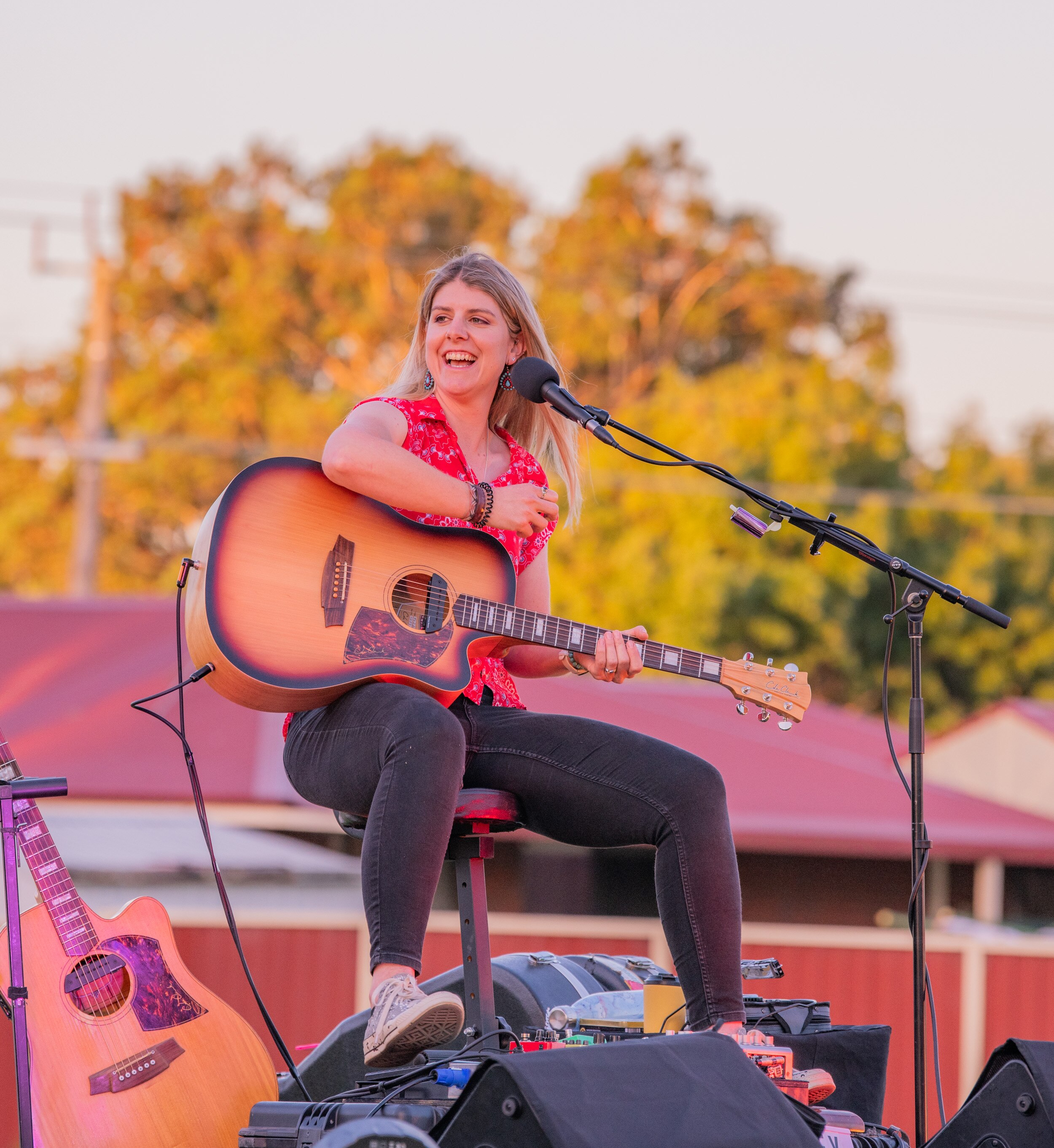 Young woman sits on stage outdoors with a guitar and microphone. She's smiling, wearing a bright red top and black jeans.