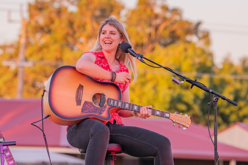 Young woman sits on stage outdoors with a guitar and microphone. She's smiling, wearing a bright red top and black jeans.
