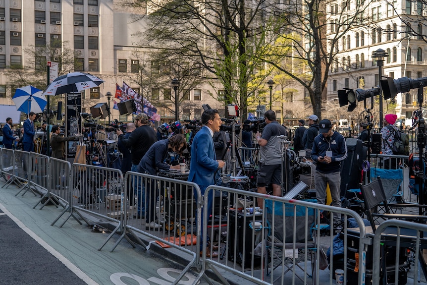Behind a metal barricade, reporters stand in a line and speak into TV cameras. Manhattan buildings are seen in the background.