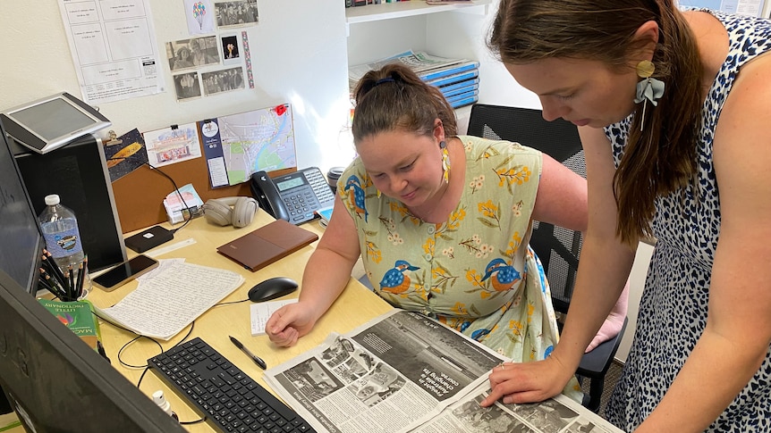 two women at a desk looking down at an open newspaper
