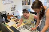Two women at a desk looking down at an open newspaper.
