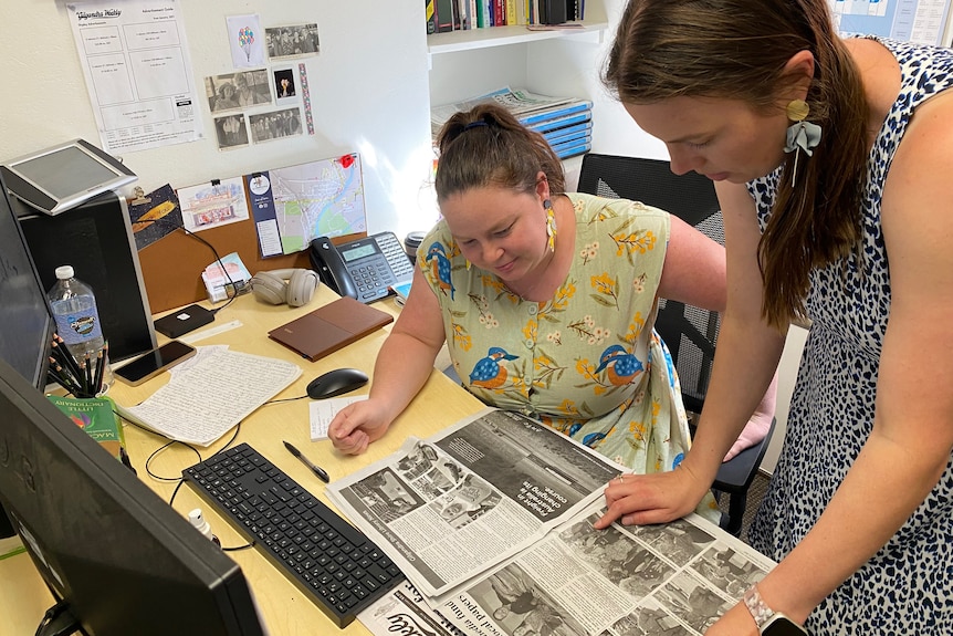 two women at a desk looking down at an open newspaper